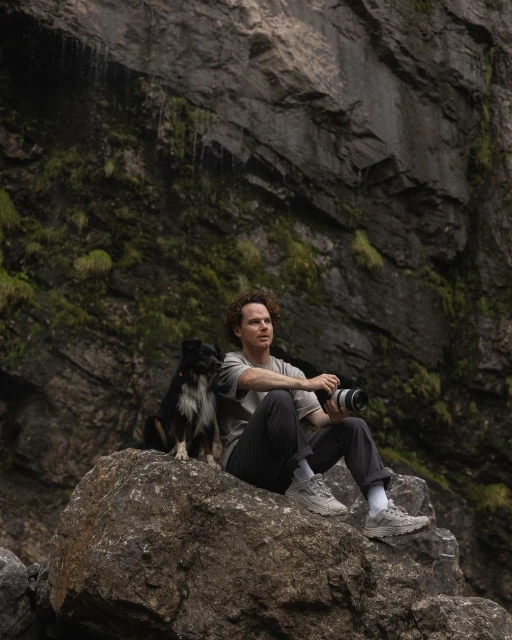 Man and dog sit on a rock in front of a rock cliff. The man holds a camera. The setting is outdoors with rocky and green foliage.