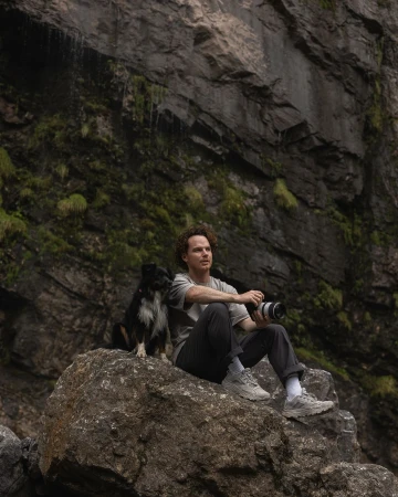 Man and dog sit on a rock in front of a rock cliff. The man holds a camera. The setting is outdoors with rocky and green foliage.