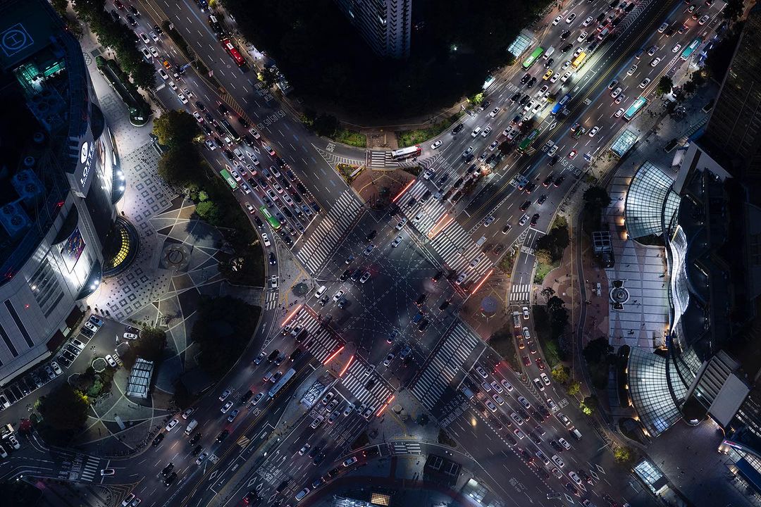 Aerial view of a bustling city intersection at night with dense traffic, illuminated buildings, and crosswalks, creating a vibrant urban scene.