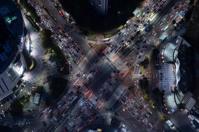 Aerial view of a bustling city intersection at night with dense traffic, illuminated buildings, and crosswalks, creating a vibrant urban scene.