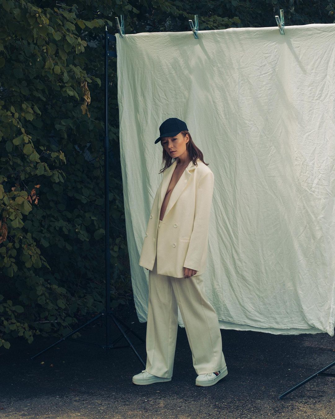 Woman in oversized white suit and baseball cap stands in front of a white sheet outdoors. Green foliage is in the background.