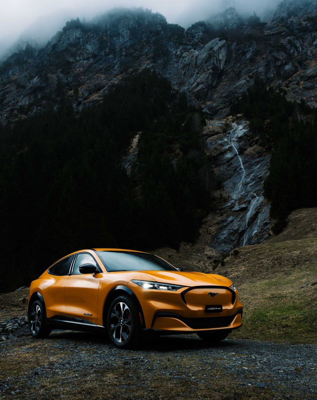 A golden yellow Ford Mustang Mach-E electric SUV is parked on a gravel road with a mountain and waterfall in the background.