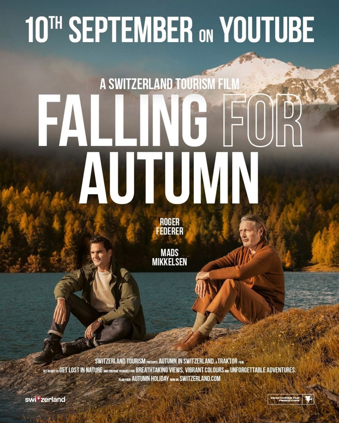 Two men, Roger Federer and Mads Mikkelsen, sit by a lake with autumn trees and a mountain in the background, promoting Switzerland tourism.