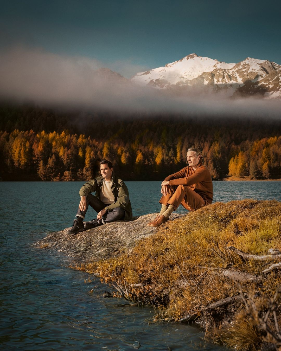 Two men in casual clothing sit on a rock near a lake surrounded by autumn trees, with snow-capped mountains in the background under a blue sky.
