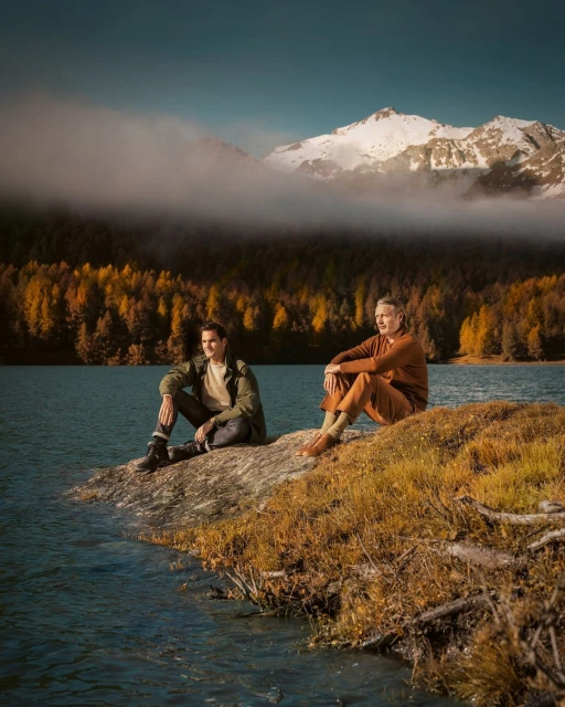 Two men in casual clothing sit on a rock near a lake surrounded by autumn trees, with snow-capped mountains in the background under a blue sky.