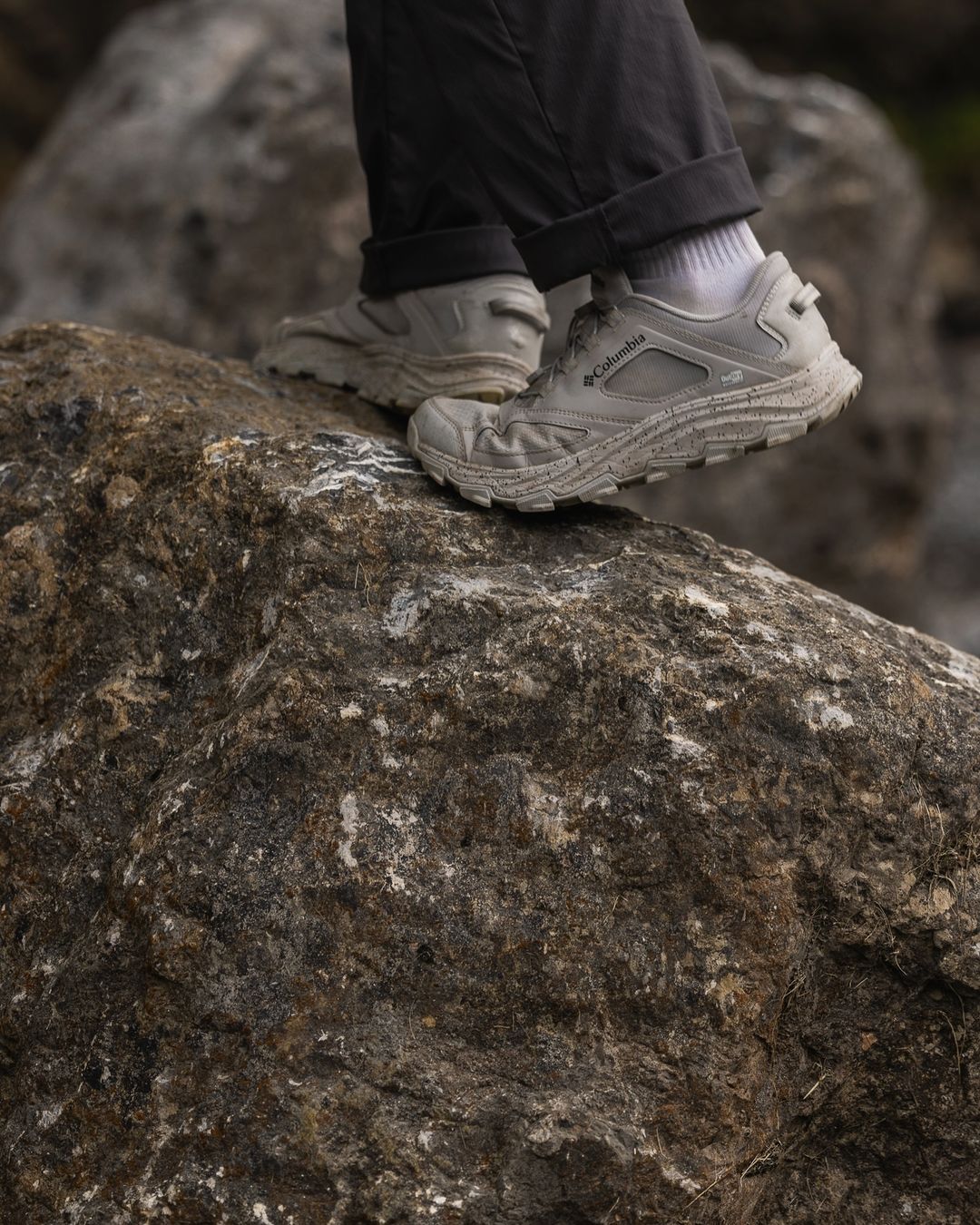 Close-up of gray Columbia hiking shoes on a textured rock surface, worn with white socks and dark gray pants in an outdoor setting.