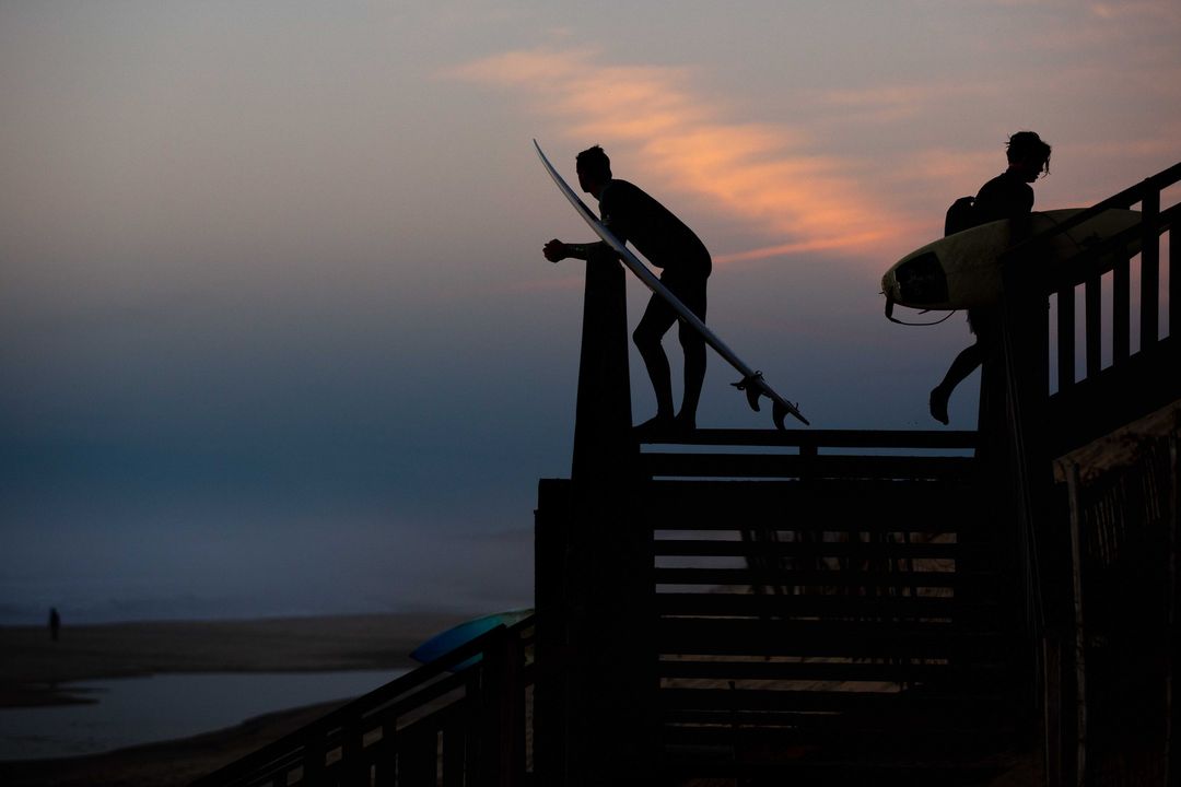 Silhouette of two surfers with surfboards on a wooden staircase against a dramatic sunset sky, creating a striking contrast.