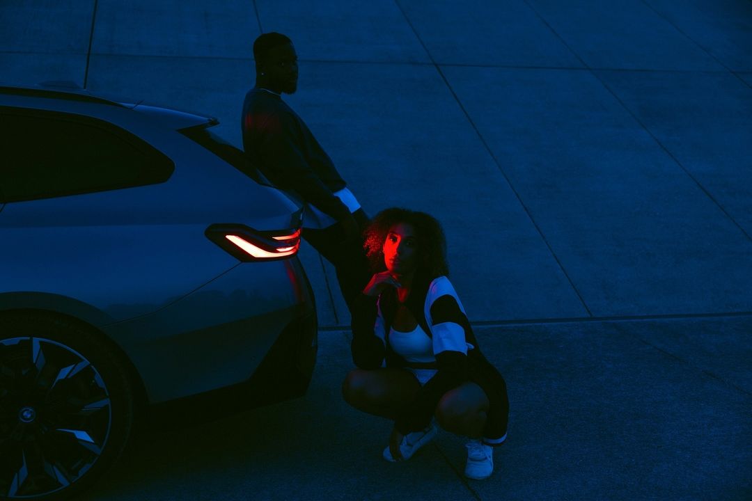 Woman crouches in front of a car as a man leans on the vehicle; both illuminated by red and blue lighting against a concrete backdrop.
