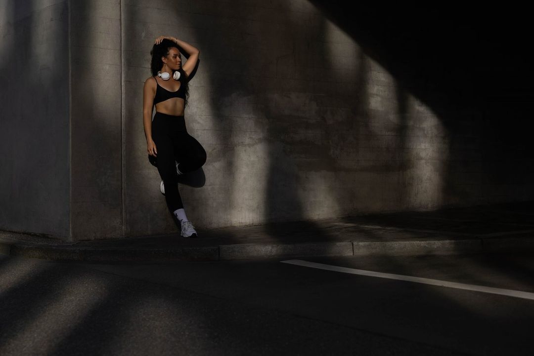 Athletic woman in black workout clothes poses against a concrete wall with her hand in her hair, wearing white headphones and sneakers.