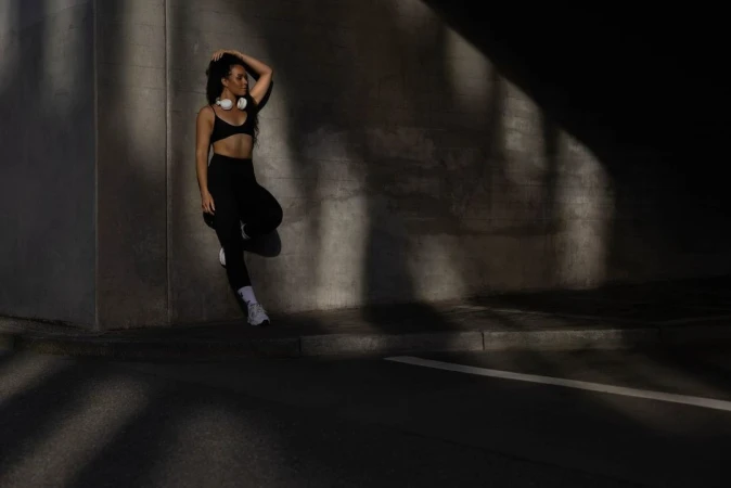 Athletic woman in black workout clothes poses against a concrete wall with her hand in her hair, wearing white headphones and sneakers.