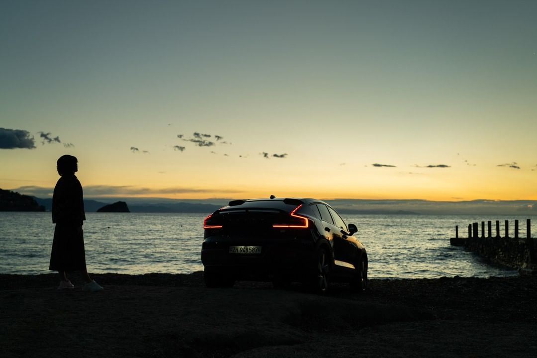 Silhouette of a person on a beach next to a black car at sunset, with calm water, a distant island, and the colorful sky.