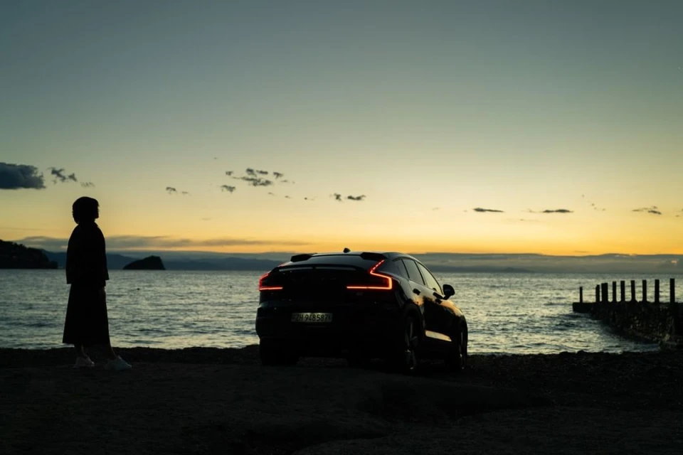 Silhouette of a person on a beach next to a black car at sunset, with calm water, a distant island, and the colorful sky.