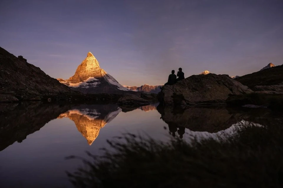 Scenic view of Matterhorn mountain reflecting in a lake at sunset, with two people sitting on a rock enjoying the view.
