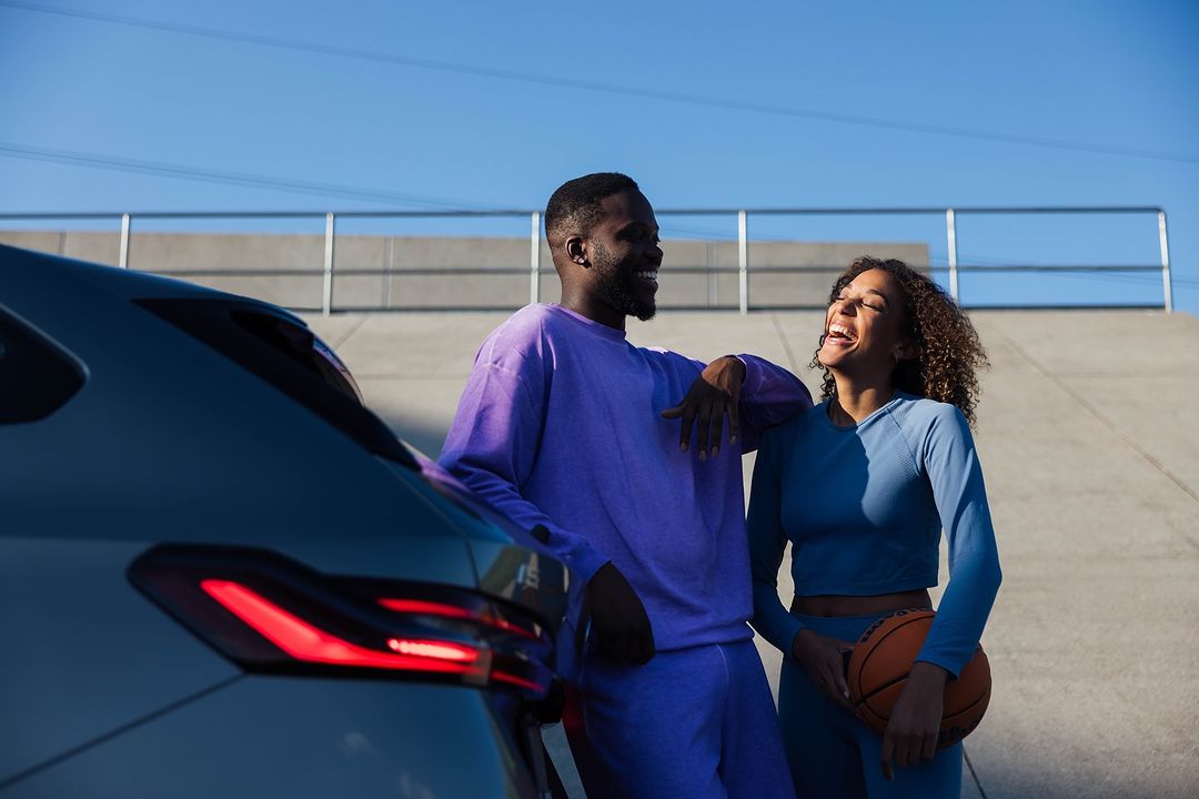 An African-American couple, dressed in athletic wear, smile while leaning against a car with a basketball on an outdoor concrete surface.