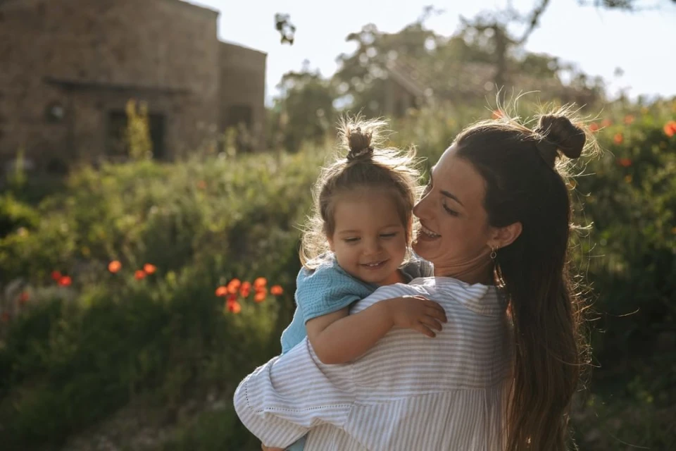 Smiling mother holding her young child in a garden setting with a building in the background, bathed in soft, natural light.