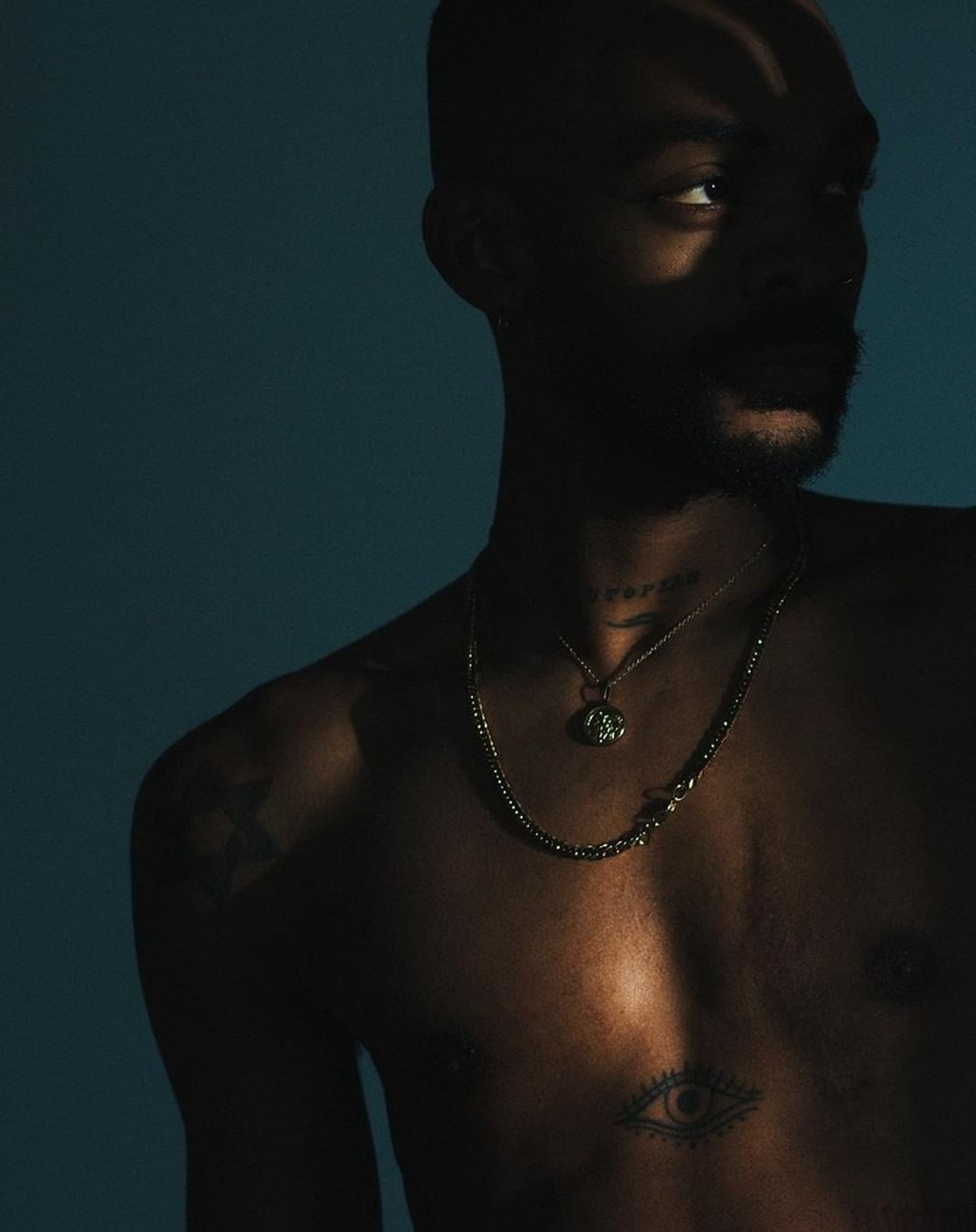 Portrait of a man with a bare chest, wearing layered necklaces in front of a dark blue background with shadows creating dramatic lighting.