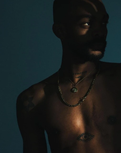 Portrait of a man with a bare chest, wearing layered necklaces in front of a dark blue background with shadows creating dramatic lighting.