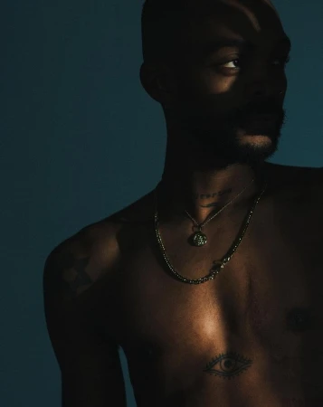 Portrait of a man with a bare chest, wearing layered necklaces in front of a dark blue background with shadows creating dramatic lighting.