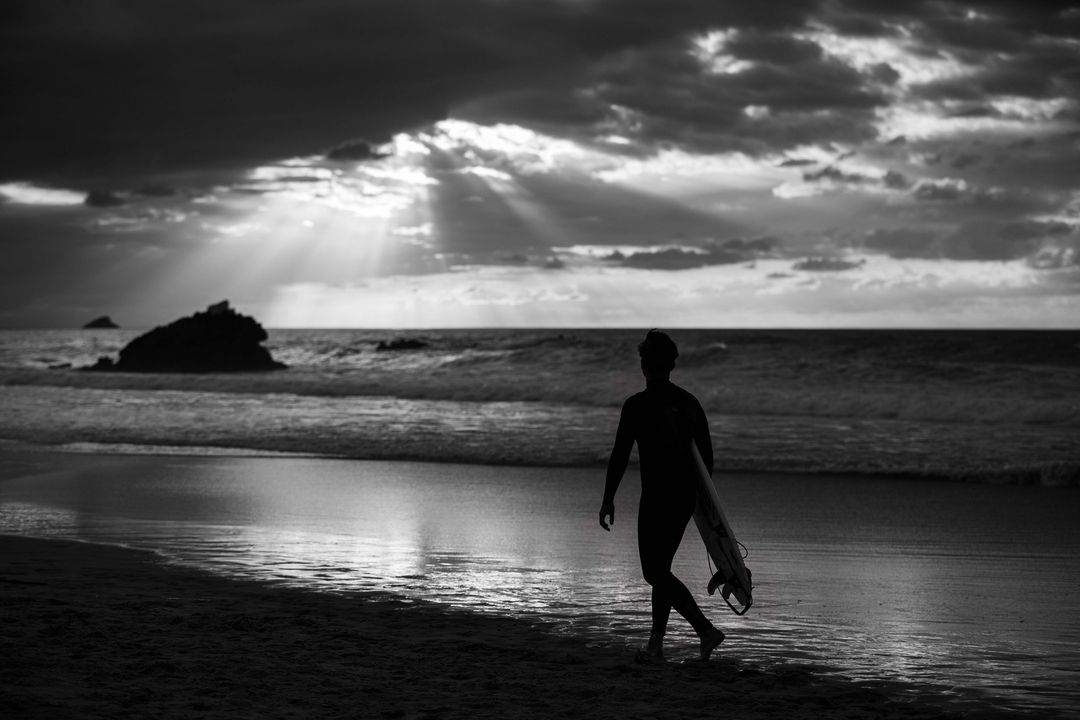 Monochrome image of a surfer walking on a beach at dusk, carrying a surfboard, with dramatic clouds and sunrays in the background.