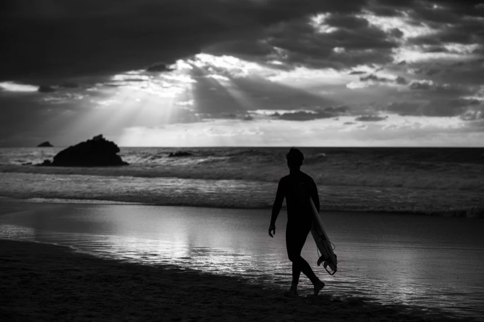 Monochrome image of a surfer walking on a beach at dusk, carrying a surfboard, with dramatic clouds and sunrays in the background.