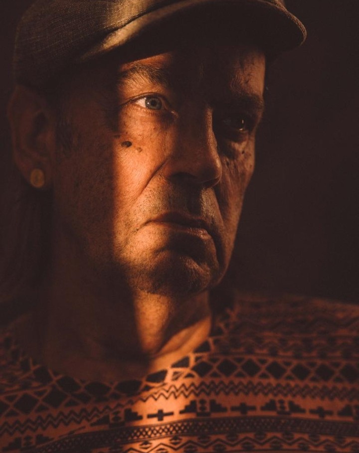 Dramatic portrait of a mature man with a cap, captured in warm, directional lighting emphasizing his face and patterned sweater indoors.