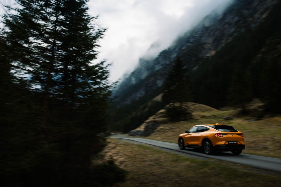 An orange Ford Mustang Mach-E drives on a winding road through a mountainous landscape with evergreen trees and fog, captured from behind.
