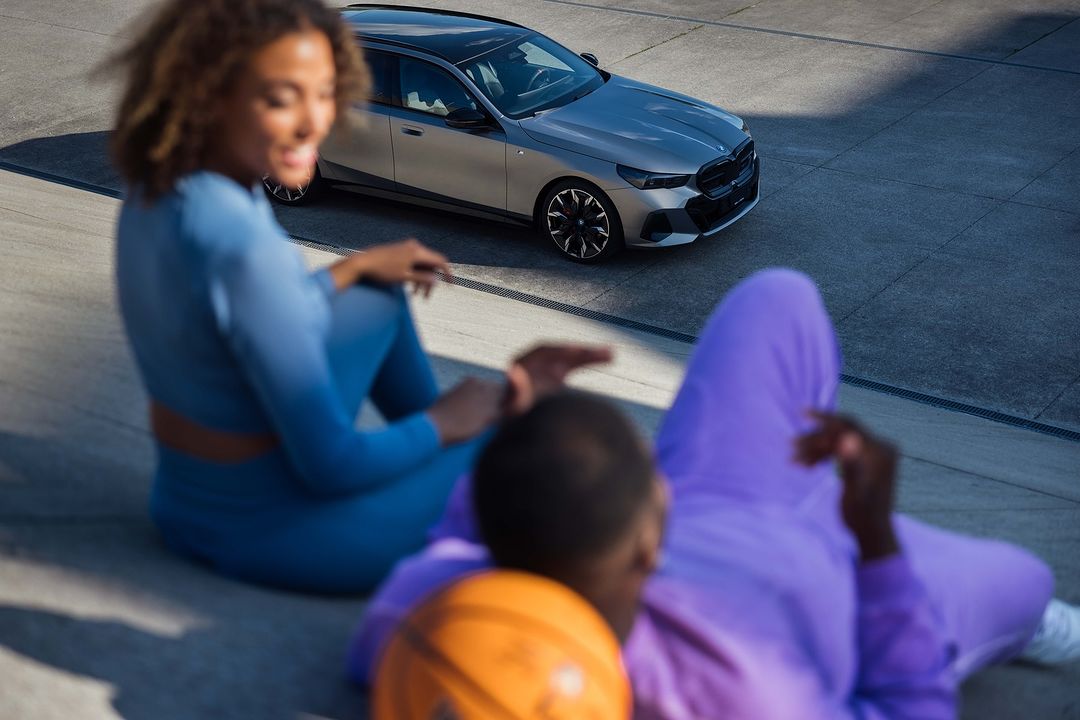Silver BMW station wagon on concrete with two adults, one holding a basketball, bathed in daylight, capturing an automotive lifestyle scene.