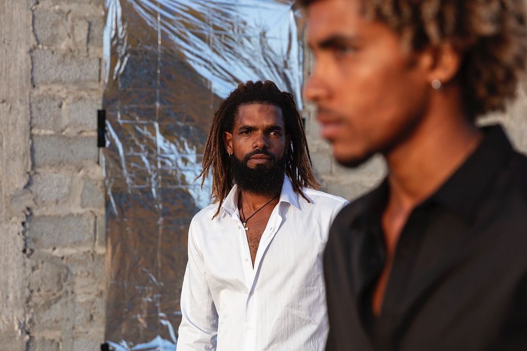 Two men pose outdoors in casual button-down shirts against a textured wall and reflective material, bathed in warm sunlight.