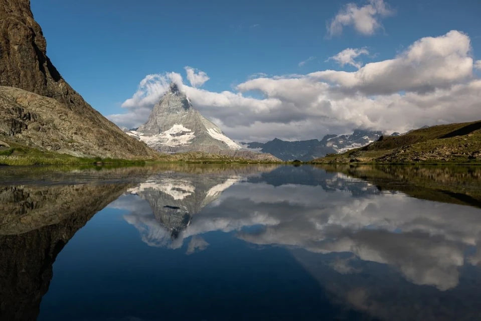 Scenic landscape featuring the Matterhorn mountain reflected in a serene lake under a partially cloudy blue sky, emphasizing natural beauty.