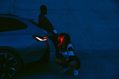 A man leaning against a car and a woman crouching near its rear light in blue lighting by Dominik Baur.