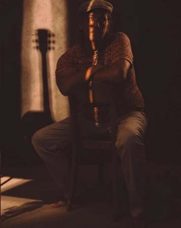 Seated man wearing a cap leaning on a wooden chair with a guitar shadow cast on the wall in a low-key portrait by Dominik Baur.