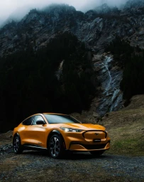 Orange electric crossover car parked on a dirt path beneath a steep rocky mountain with a visible waterfall.
