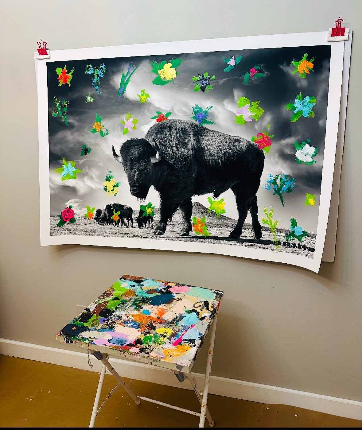 Artwork featuring a bison and painted flowers, displayed indoors on a wall, with a paint-covered folding table in the foreground.