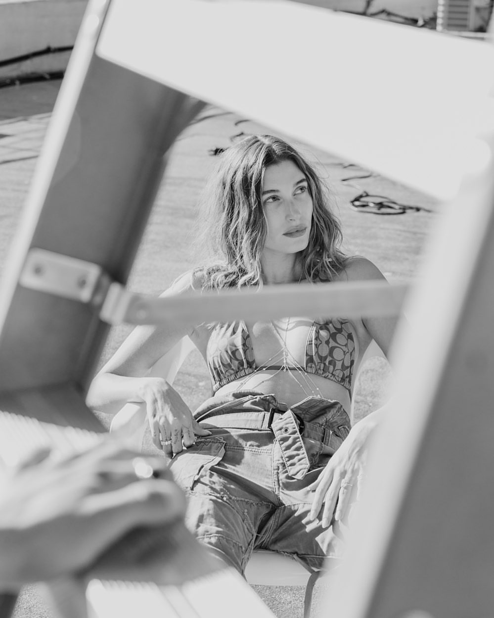 Black and white portrait of a woman with wavy hair, wearing a patterned top and high-waisted belted shorts, framed by a ladder.