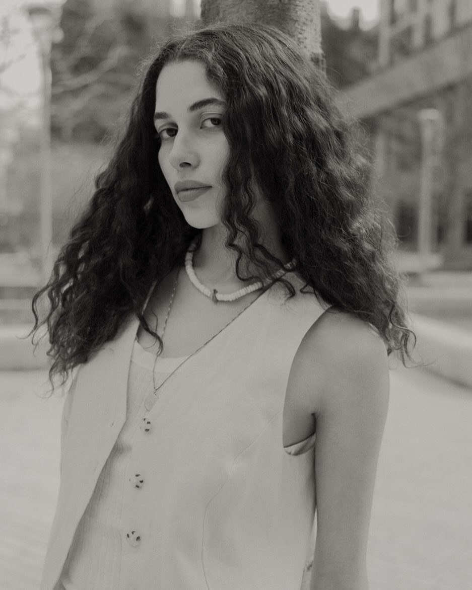 Monochrome portrait of a woman with curly hair wearing a vest and necklace standing outdoors with trees in the background.