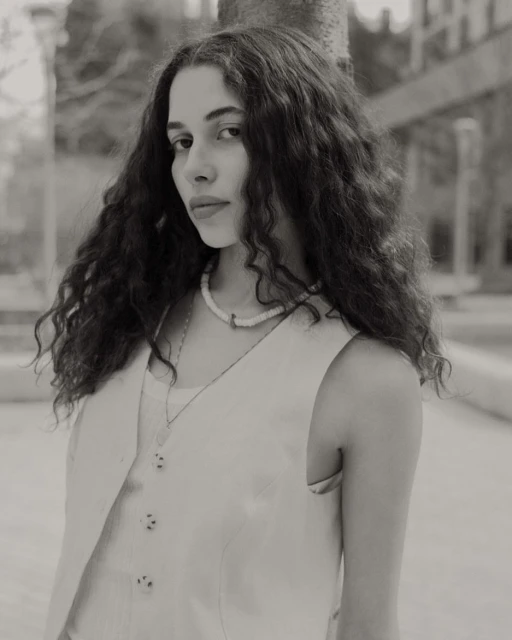 Monochrome portrait of a woman with curly hair wearing a vest and necklace standing outdoors with trees in the background.