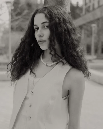 Monochrome portrait of a woman with curly hair wearing a vest and necklace standing outdoors with trees in the background.
