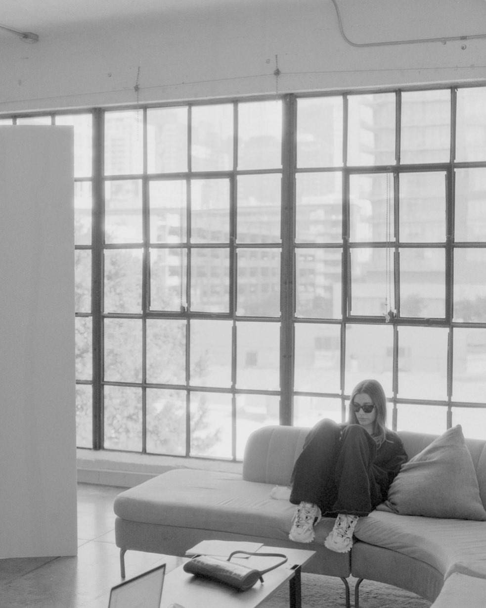 Monochrome shot of an adult woman in sunglasses, casually posed on a sofa in a bright loft apartment by a large window.