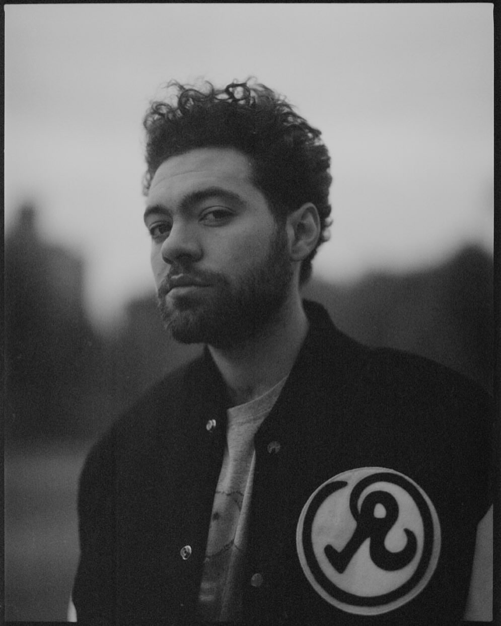 Black and white portrait of a man with curly hair wearing a letterman jacket with a circular patch, set against a blurred outdoor background.