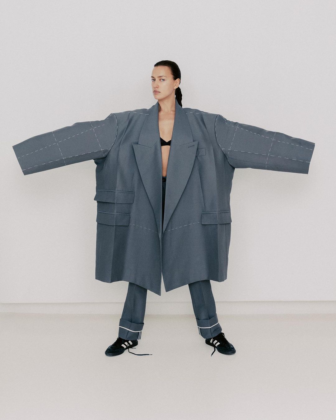 Fashion portrait of a woman wearing an oversized grey coat and matching pants with black sneakers against a neutral white background.