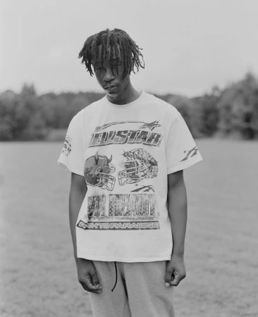Monochrome portrait of a young Black man standing outdoors wearing a graphic t-shirt and light trousers.