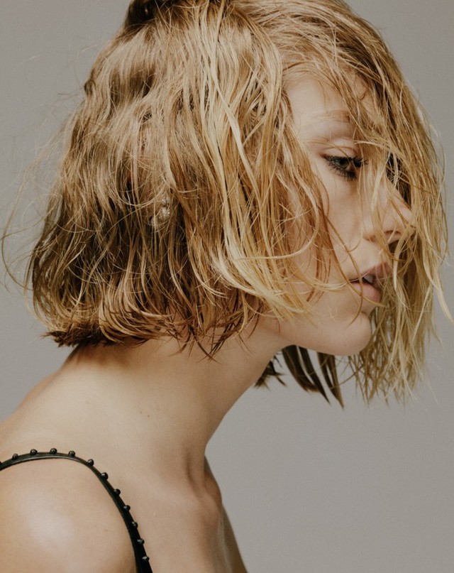 Close-up portrait of a fair-skinned woman with wet, blonde, bobbed hair, gazing slightly downward, wearing a black top against a gray background.