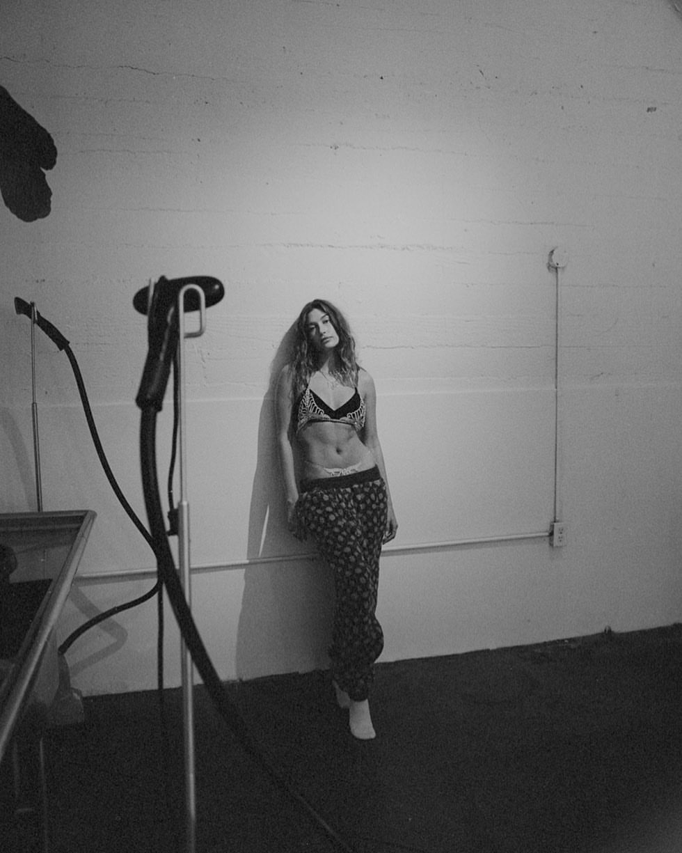 Woman poses against white wall in photo studio wearing a bra top and pajama pants. Steam iron and surface visible.