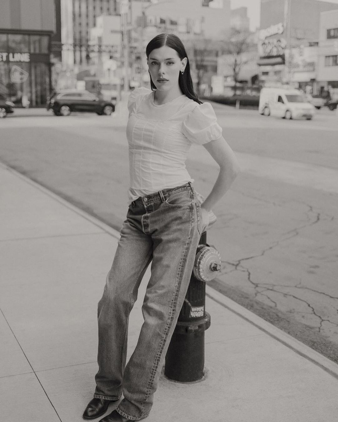 Woman in jeans and white blouse poses on urban street next to fire hydrant, with city buildings and traffic in the background.
