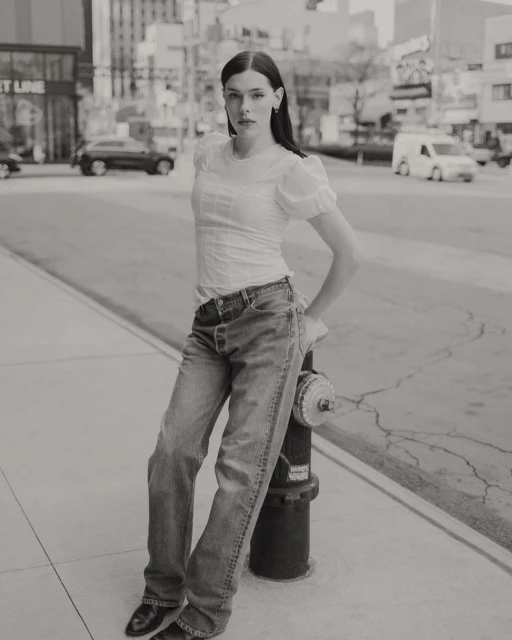 Woman in jeans and white blouse poses on urban street next to fire hydrant, with city buildings and traffic in the background.
