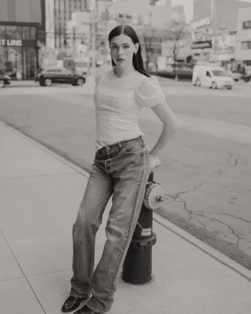 Woman in jeans and white blouse poses on urban street next to fire hydrant, with city buildings and traffic in the background.