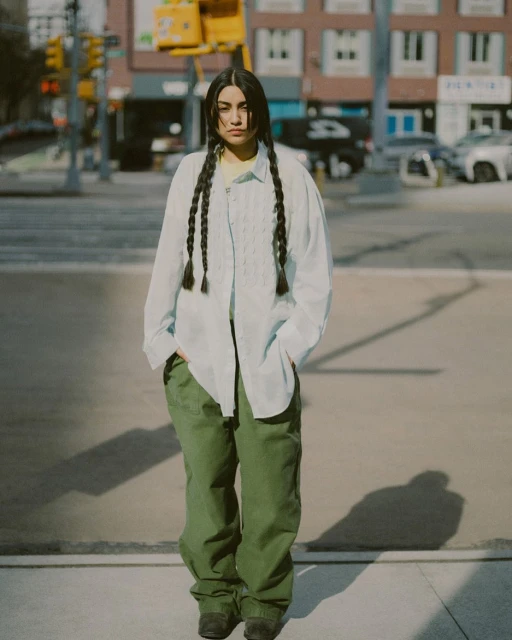 Portrait of a young woman with long dark braids, posing in an open light blue shirt and olive pants, standing on a city street.