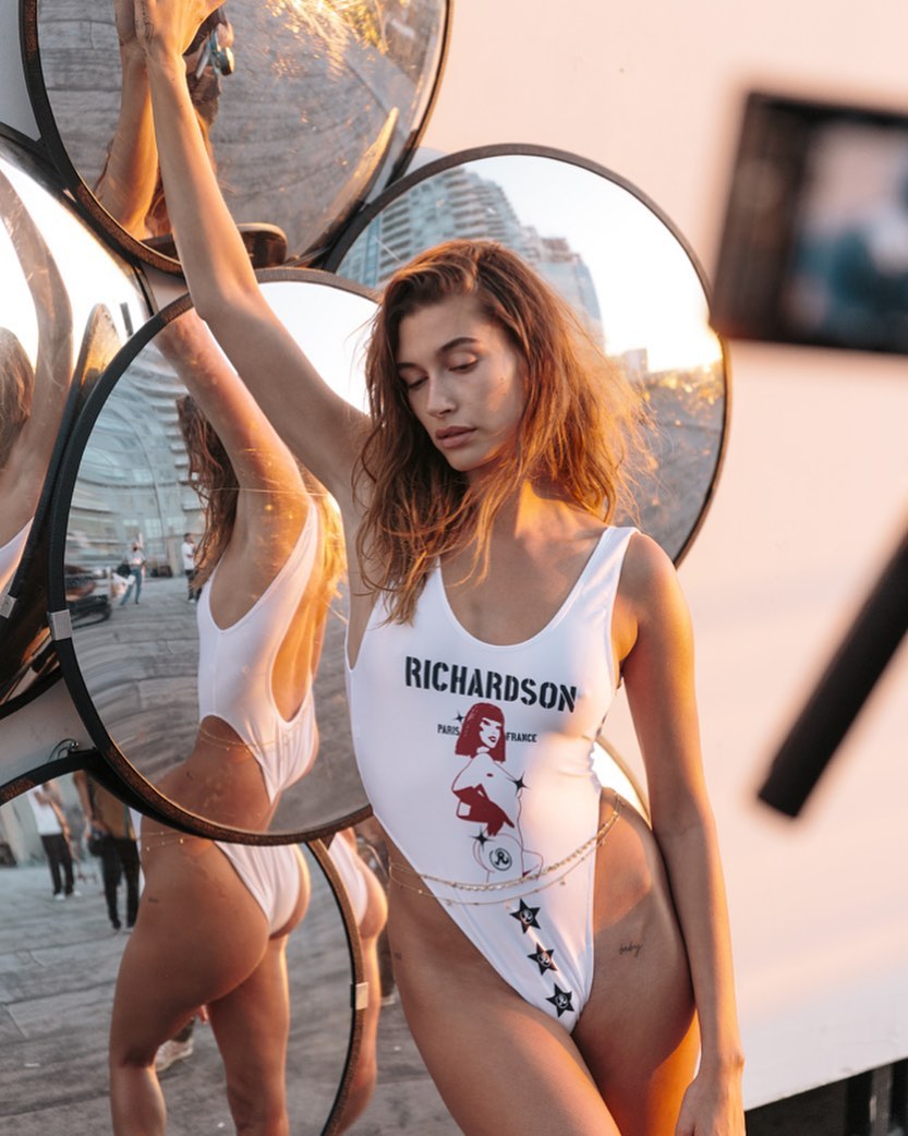 Adult female fashion model poses in a white swimsuit with a graphic design, with multiple round mirrors in an outdoor environment.