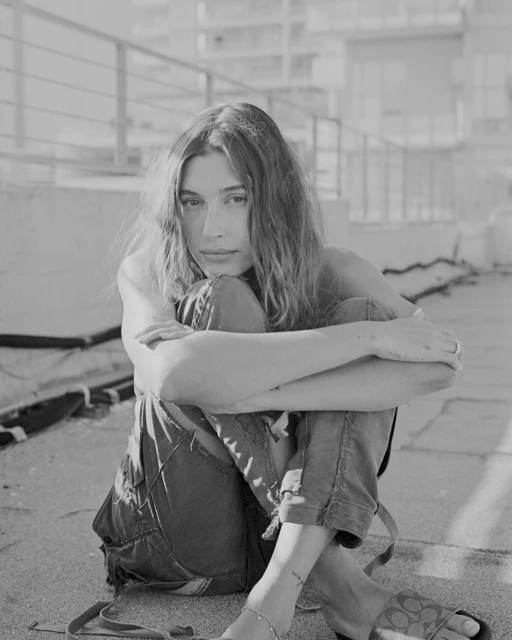 Black and white portrait of a woman with long hair, wearing overalls and sandals, sitting outdoors with her arms around her knees.
