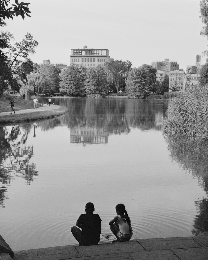 Black and white shot of two people sitting by a lake with buildings and trees in the background, reflecting in the water.